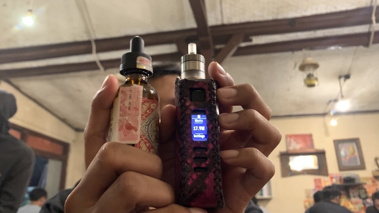 A person is holding a vaping device in their right hand and a bottle of e-liquid in their left hand. The background features an indoor setting with visible wooden ceiling beams, some lit hanging lights, wall decorations including pictures or posters, and other people present.