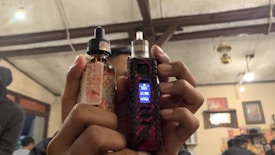 A person is holding a vaping device in their right hand and a bottle of e-liquid in their left hand. The background features an indoor setting with visible wooden ceiling beams, some lit hanging lights, wall decorations including pictures or posters, and other people present.