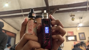 A person is holding a vaping device in their right hand and a bottle of e-liquid in their left hand. The background features an indoor setting with visible wooden ceiling beams, some lit hanging lights, wall decorations including pictures or posters, and other people present.