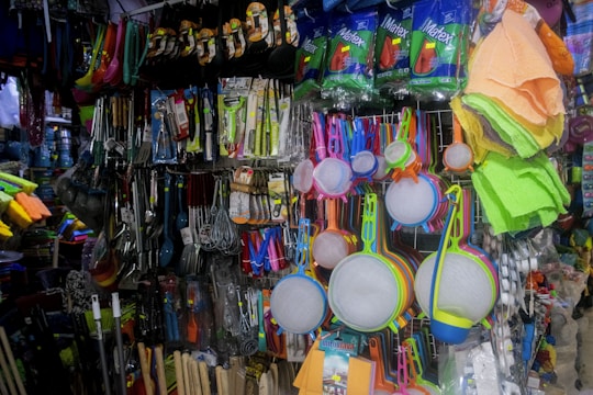 A variety of kitchen utensils and cleaning supplies are displayed on a densely packed store shelf. The items include colorful strainers, sponges, brooms, and packaged kitchen tools like spatulas and ladles. The display is vibrant with a mix of colors and textures.