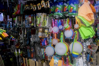 A variety of kitchen utensils and cleaning supplies are displayed on a densely packed store shelf. The items include colorful strainers, sponges, brooms, and packaged kitchen tools like spatulas and ladles. The display is vibrant with a mix of colors and textures.