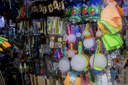 A variety of kitchen utensils and cleaning supplies are displayed on a densely packed store shelf. The items include colorful strainers, sponges, brooms, and packaged kitchen tools like spatulas and ladles. The display is vibrant with a mix of colors and textures.