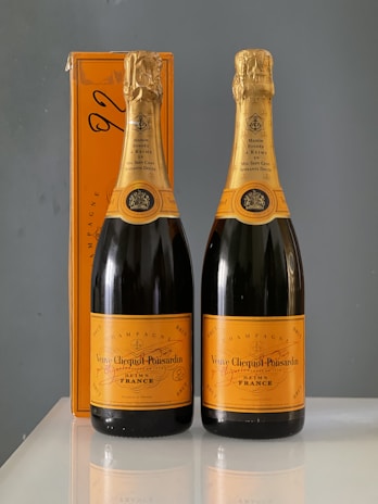 Two bottles of Veuve Clicquot Champagne are placed side by side on a reflective surface. They feature signature orange labels and gold foil around the necks. An orange box designed to match the bottles is positioned behind them.