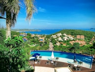 A panoramic view of a lush green landscape overlooking a coastal area. In the foreground, there is a luxurious infinity pool surrounded by chairs and umbrellas. Beyond the pool, a road extends through the greenery leading to a residential area with various houses and buildings featuring terracotta roofs. The vibrant blue sea stretches across the horizon under a clear sky, with a distant island visible on the right.
