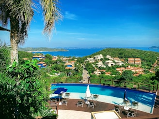 A panoramic view of a lush green landscape overlooking a coastal area. In the foreground, there is a luxurious infinity pool surrounded by chairs and umbrellas. Beyond the pool, a road extends through the greenery leading to a residential area with various houses and buildings featuring terracotta roofs. The vibrant blue sea stretches across the horizon under a clear sky, with a distant island visible on the right.