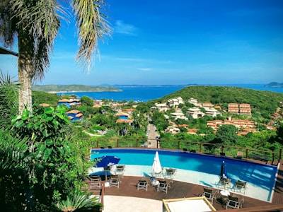 A panoramic view of a lush green landscape overlooking a coastal area. In the foreground, there is a luxurious infinity pool surrounded by chairs and umbrellas. Beyond the pool, a road extends through the greenery leading to a residential area with various houses and buildings featuring terracotta roofs. The vibrant blue sea stretches across the horizon under a clear sky, with a distant island visible on the right.