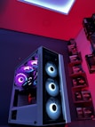A gaming computer setup featuring a high-performance PC tower with multiple RGB fans lit in blue and purple hues. The case has a modern design with transparent panels showcasing internal components. In the background, shelves are stocked with various Ryzen and other gaming components under a vibrant red and blue ambient light.
