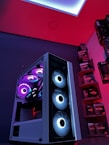 A gaming computer setup featuring a high-performance PC tower with multiple RGB fans lit in blue and purple hues. The case has a modern design with transparent panels showcasing internal components. In the background, shelves are stocked with various Ryzen and other gaming components under a vibrant red and blue ambient light.