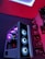 A gaming computer setup featuring a high-performance PC tower with multiple RGB fans lit in blue and purple hues. The case has a modern design with transparent panels showcasing internal components. In the background, shelves are stocked with various Ryzen and other gaming components under a vibrant red and blue ambient light.