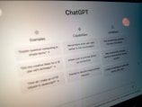 A close-up view of a computer screen displaying the ChatGPT interface. The screen is divided into three sections labeled 'Examples,' 'Capabilities,' and 'Limitations,' each containing text descriptions. The environment appears dimly lit, emphasizing the screen's brightness and focus.