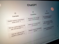 A close-up view of a computer screen displaying the ChatGPT interface. The screen is divided into three sections labeled 'Examples,' 'Capabilities,' and 'Limitations,' each containing text descriptions. The environment appears dimly lit, emphasizing the screen's brightness and focus.