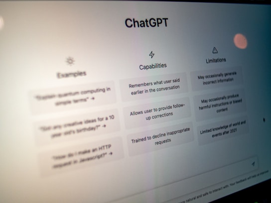 A close-up view of a computer screen displaying the ChatGPT interface. The screen is divided into three sections labeled 'Examples,' 'Capabilities,' and 'Limitations,' each containing text descriptions. The environment appears dimly lit, emphasizing the screen's brightness and focus.
