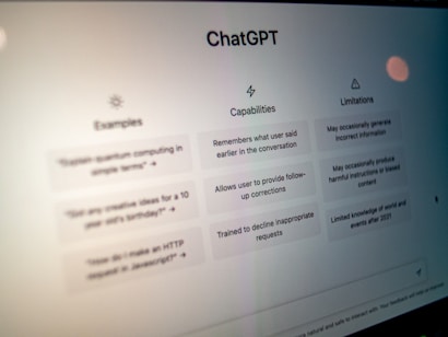 A close-up view of a computer screen displaying the ChatGPT interface. The screen is divided into three sections labeled 'Examples,' 'Capabilities,' and 'Limitations,' each containing text descriptions. The environment appears dimly lit, emphasizing the screen's brightness and focus.