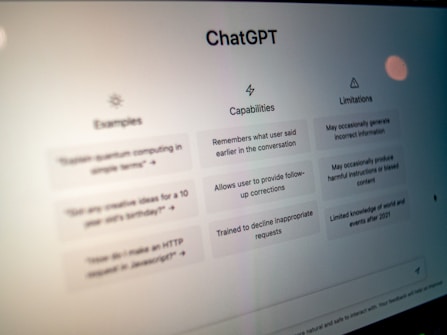 A close-up view of a computer screen displaying the ChatGPT interface. The screen is divided into three sections labeled 'Examples,' 'Capabilities,' and 'Limitations,' each containing text descriptions. The environment appears dimly lit, emphasizing the screen's brightness and focus.