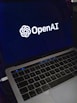 A laptop screen displaying the OpenAI logo and text. The laptop keyboard is visible below, with keys illuminated in a dimly lit environment.