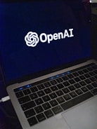 A laptop screen displaying the OpenAI logo and text. The laptop keyboard is visible below, with keys illuminated in a dimly lit environment.
