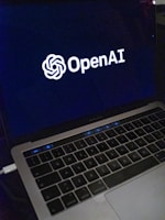 A laptop screen displaying the OpenAI logo and text. The laptop keyboard is visible below, with keys illuminated in a dimly lit environment.