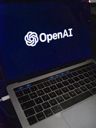 A laptop screen displaying the OpenAI logo and text. The laptop keyboard is visible below, with keys illuminated in a dimly lit environment.