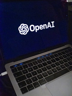 A laptop screen displaying the OpenAI logo and text. The laptop keyboard is visible below, with keys illuminated in a dimly lit environment.