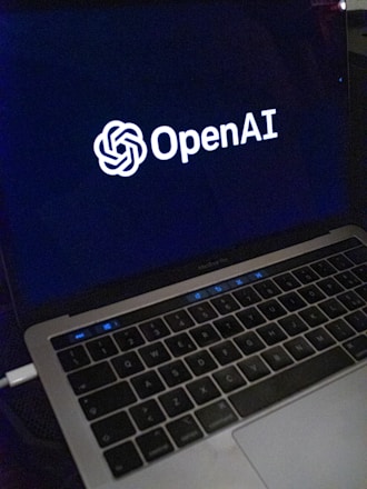 A laptop screen displaying the OpenAI logo and text. The laptop keyboard is visible below, with keys illuminated in a dimly lit environment.