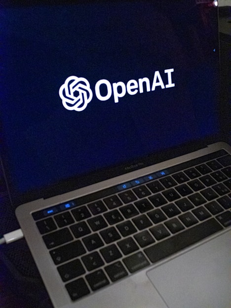 A laptop screen displaying the OpenAI logo and text. The laptop keyboard is visible below, with keys illuminated in a dimly lit environment.
