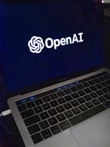 A laptop screen displaying the OpenAI logo and text. The laptop keyboard is visible below, with keys illuminated in a dimly lit environment.