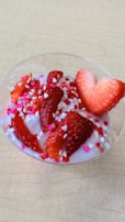 A bowl of yogurt decorated with sliced strawberries and small heart-shaped sprinkles. The yogurt appears to be a light pink color, complementing the red of the strawberries. The sprinkles add a whimsical touch with colors in shades of pink and white, enhancing the visual appeal.