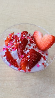 A bowl of yogurt decorated with sliced strawberries and small heart-shaped sprinkles. The yogurt appears to be a light pink color, complementing the red of the strawberries. The sprinkles add a whimsical touch with colors in shades of pink and white, enhancing the visual appeal.