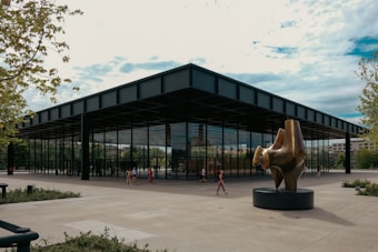 A modernist-style building with large glass walls and a flat roof is surrounded by a spacious plaza. Several people are casually walking around, providing a sense of scale to the minimalist architecture. In the foreground, there is a large abstract bronze sculpture placed on a pedestal. Lush greenery and trees touch the scene on the sides, blending natural elements with the urban setting.