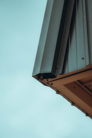 An architectural detail of a building featuring metal cladding with a minimalist design approach. The metal panels are gently angled and appear weathered with small spots visible on the surface.