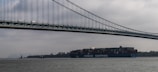 A large cargo ship labeled 'COSCO Shipping' is moving underneath a towering suspension bridge. The sky is overcast, creating a moody atmosphere. In the background, several small boats and a foggy city skyline enhance the scene.