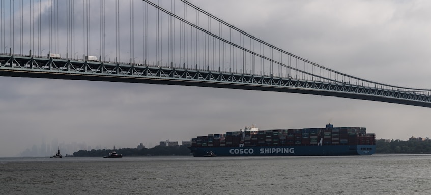A large cargo ship labeled 'COSCO Shipping' is moving underneath a towering suspension bridge. The sky is overcast, creating a moody atmosphere. In the background, several small boats and a foggy city skyline enhance the scene.