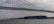 A large cargo ship labeled 'COSCO Shipping' is moving underneath a towering suspension bridge. The sky is overcast, creating a moody atmosphere. In the background, several small boats and a foggy city skyline enhance the scene.
