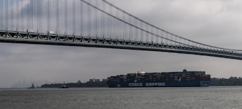 A large cargo ship labeled 'COSCO Shipping' is moving underneath a towering suspension bridge. The sky is overcast, creating a moody atmosphere. In the background, several small boats and a foggy city skyline enhance the scene.