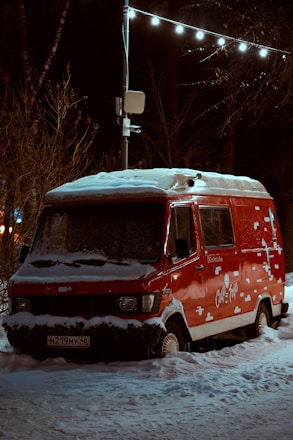A red van covered in snow is parked against a backdrop of bare trees at night. Warm light from street lamps illuminates the scene, highlighting the snow on the vehicle roof and around the wheels. The van displays decorative white graphics resembling clouds or snowflakes.