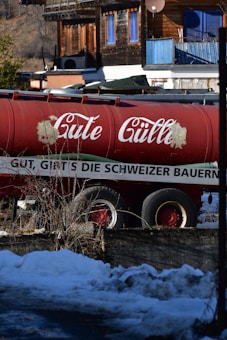 A large red tanker truck is parked in front of a rustic wooden building with blue shutters. The tanker has white text on its side. Snow and small bushes are visible in the foreground, indicating a cold environment.