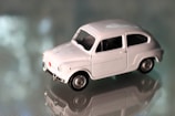 A small white toy car with detailed features sits on a reflective surface. The background is blurred, highlighting the sleek design and vintage style of the toy model.
