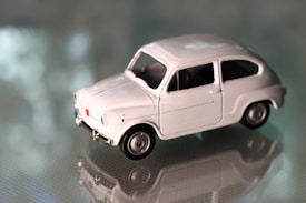 A small white toy car with detailed features sits on a reflective surface. The background is blurred, highlighting the sleek design and vintage style of the toy model.