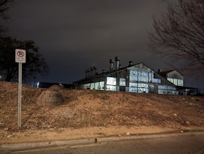 A dimly lit industrial building with 'Marc Steel Company' written on it, featuring large windows reflecting light. It sits behind a barren, sloped area with a no parking sign. Bare, leafless trees frame the scene, conveying a sense of neglect or abandonment. The sky is overcast, enhancing the moody atmosphere.