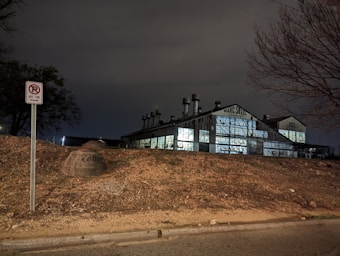 A dimly lit industrial building with 'Marc Steel Company' written on it, featuring large windows reflecting light. It sits behind a barren, sloped area with a no parking sign. Bare, leafless trees frame the scene, conveying a sense of neglect or abandonment. The sky is overcast, enhancing the moody atmosphere.