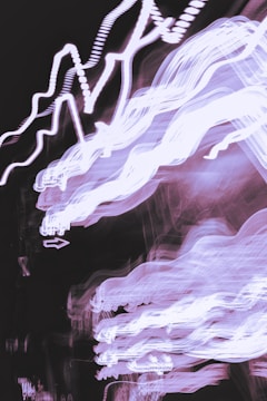 Abstract light trails in white and purple tones create a dynamic and chaotic pattern against a dark background. The lines appear jagged and fluid, resembling waves or electrical pulses.