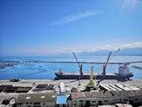 A large cargo ship docked at a port with two cranes positioned over it. The sky is clear and blue with scattered clouds, and the sea is calm. There are mountain ranges visible in the background across the water. Various containers and industrial buildings are present on the dock.