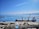 A large cargo ship docked at a port with two cranes positioned over it. The sky is clear and blue with scattered clouds, and the sea is calm. There are mountain ranges visible in the background across the water. Various containers and industrial buildings are present on the dock.