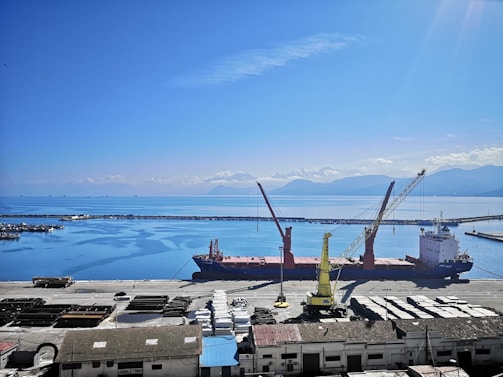 A large cargo ship docked at a port with two cranes positioned over it. The sky is clear and blue with scattered clouds, and the sea is calm. There are mountain ranges visible in the background across the water. Various containers and industrial buildings are present on the dock.