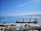 A large cargo ship docked at a port with two cranes positioned over it. The sky is clear and blue with scattered clouds, and the sea is calm. There are mountain ranges visible in the background across the water. Various containers and industrial buildings are present on the dock.