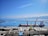 A large cargo ship docked at a port with two cranes positioned over it. The sky is clear and blue with scattered clouds, and the sea is calm. There are mountain ranges visible in the background across the water. Various containers and industrial buildings are present on the dock.