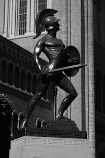 A bronze statue of a warrior in classical attire stands on a pedestal, holding a sword and shield. Positioned in a heroic and dynamic pose, the statue captures the strength and valor typically associated with ancient warriors. The architecture in the background suggests a classical or academic setting, with brick walls and tall windows.