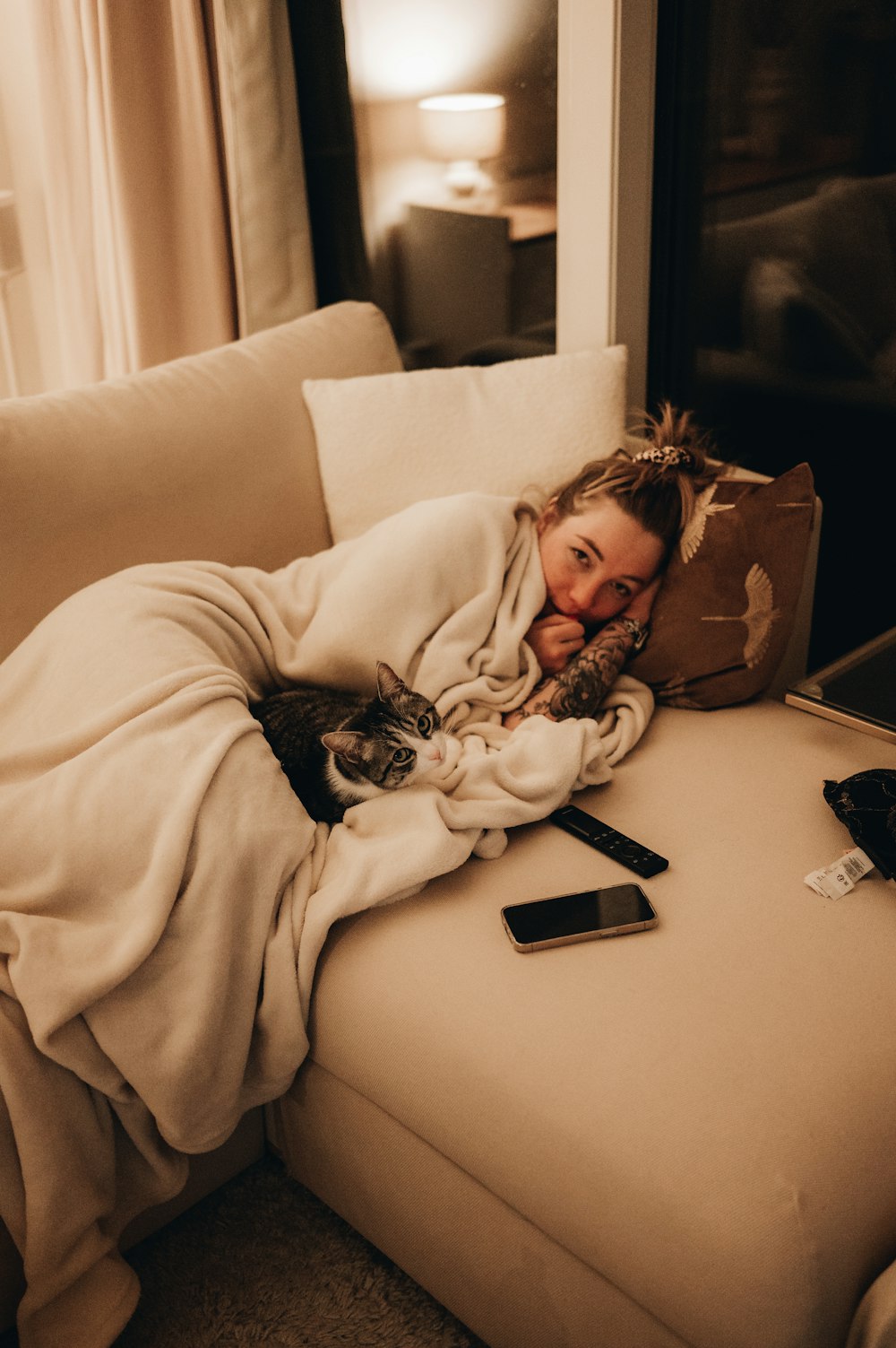 a woman laying on a couch with a cat under a blanket