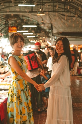 Two women smiling and holding hands in a crowded market setting. The woman on the left wears a colorful dress with a lemon pattern, and the woman on the right is in a white dress. The background is busy with people and various lights around.