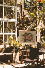 A sunlit garden area features several potted plants and a framed painting on an easel depicting pink flowers. The scene is set against a backdrop of a glass window with vine-covered walls. A foldable chair is placed in the foreground, and various gardening tools are scattered around.